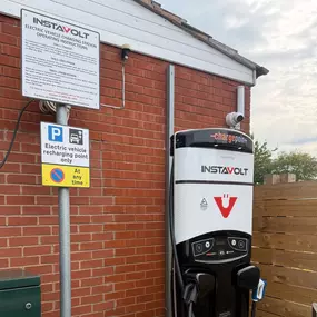 A close-up of an InstaVolt charger installed beside a red brick building, featuring CCTV and signage for EV drivers, including 24-hour parking restrictions.