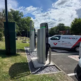 Side view of InstaVolt EV chargers in use, with an InstaVolt-branded electric van parked nearby and picnic benches visible behind.