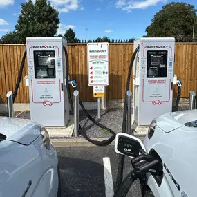 This image shows two InstaVolt ultra-rapid electric vehicle chargers in operation at The Avenue Car Park, Blakedown. Both bays are occupied by EVs, with clear signage providing user instructions. A wooden fence and trees surround the location, creating a tidy and accessible charging spot.