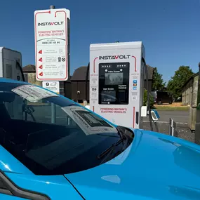 A bright blue electric vehicle is actively charging at the InstaVolt station, set in a rural environment with an old-style streetlamp and lush greenery in the background.