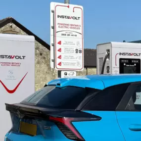 The front of a charging electric car is shown beside a close-up of the InstaVolt charger, highlighting signage and the scenic, barn-style background architecture.
