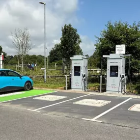 This wide-angle shot showcases multiple InstaVolt ultra rapid chargers at the Wincanton A303 site, with a blue EV actively charging. The site includes several clearly marked charging bays, surrounded by greenery and adjacent to the A303 for convenient access.