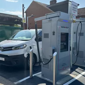 This image shows a pair of InstaVolt chargers actively in use, positioned near a brick building and a Co-operative Food store. A branded Toyota van and a green EV are parked and charging.
