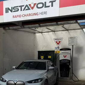 A close-up image of a silver BMW EV charging at an InstaVolt rapid charger in Braunton. The charger is installed within a sheltered bay, featuring InstaVolt branding and safety signage. The setup is clean and modern, designed for quick EV charging access alongside the main service station amenities.