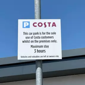 A close-up of Costa’s car park signage outlining parking terms for EV drivers and customers. It clearly states a three-hour maximum stay, only for Costa patrons, with a blue sky in the background. This image is useful for clarifying on-site parking conditions.
