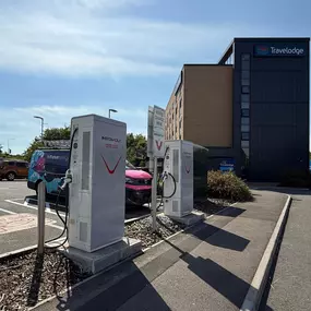 This image shows two InstaVolt ultra rapid electric vehicle chargers positioned in front of a Travelodge hotel building. An Octopus Energy van charging in the background. The scene is bright and clear, ideal for showcasing charger placement near accommodation.