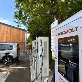A side view of InstaVolt ultra rapid chargers beside a timber-clad café building, with an EV charging under leafy trees at Three Trees Farm Shop.
