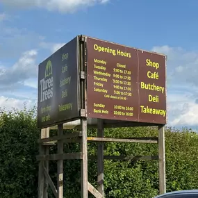 A large outdoor sign displaying the shop, café, and deli opening times for Three Trees Farm Shop, helpful for EV drivers planning a visit.