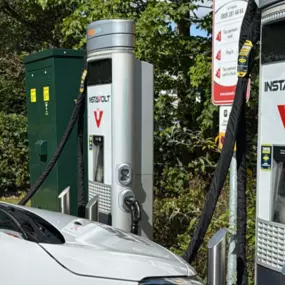 A white electric van is plugged into one of two InstaVolt rapid chargers in Shirley. The chargers are set in a green roadside location with visible signage and safety bollards.