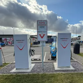 Rear view of two InstaVolt chargers at Rugeley’s Leathermill Lane site, with a blue EV parked and KFC visible behind the chargers.