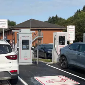 Electric vehicles using InstaVolt ultra-rapid chargers beside a red-brick facility with trees and blue skies overhead.