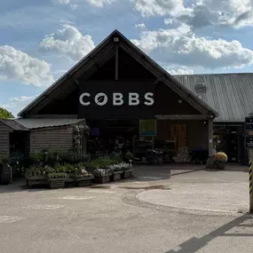 The entrance of Cobbs Farm Shop in Hungerford, located just off the A4 Bath Road. The building features a pitched roof with the Cobbs sign prominently displayed. Outdoor crates of plants and flowers enhance the welcoming countryside feel.