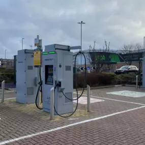 Four InstaVolt ultra-rapid chargers are lined up in a car park, with marked bays and visible retail units nearby, offering accessible charging for EV drivers.