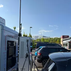 This image shows a row of InstaVolt electric vehicle chargers in operation at a retail car park in Solstice Park, Amesbury. A Costa Coffee Drive Thru is prominently visible on the right, with several EVs actively charging. In the distance, the signage for Home Bargains and other large-format stores lines the horizon, indicating the site’s convenient proximity to popular retail outlets.