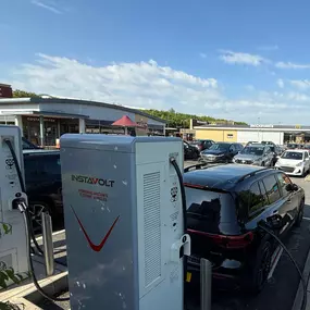 A broad view of the vehicle entrance to the Costa Coffee Drive Thru at Solstice Park, Amesbury, where InstaVolt EV chargers are located. The access road curves through a landscaped car park filled with vehicles, with directional signage guiding drivers. The presence of Costa Coffee and nearby retail outlets highlights the convenient amenities available to drivers while charging their electric vehicles with InstaVolt.