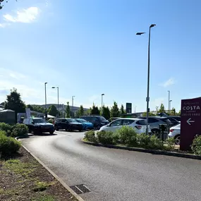 A broad view of the vehicle entrance to the Costa Coffee Drive Thru at Solstice Park, Amesbury. The access road curves through a landscaped car park with tall signage and lampposts. Numerous parked vehicles are visible, and in the background, signage for Costa and other retailers confirms the location as a popular and easily accessible destination for travellers and EV drivers alike.
