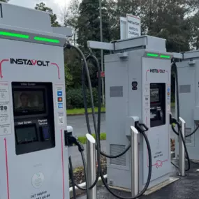 Four InstaVolt chargers positioned in a row, with one red EV in use. Each bay is clearly marked for electric vehicle charging, set beside a quiet road with trees in the background.