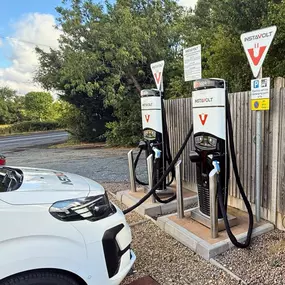 A white EV charges at one of the InstaVolt rapid charging units at Alcester Road Service Station. The location is positioned beside the A422, with rural scenery and trees in the background, offering easy roadside access.
