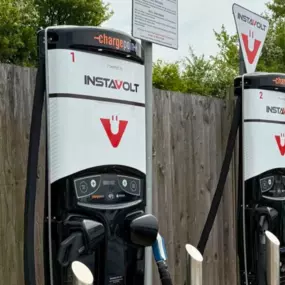 A pair of InstaVolt rapid electric vehicle chargers located at Alcester Road Service Station. Signage includes usage instructions and dedicated EV parking. The chargers are backed by a wooden fence and surrounded by greenery.