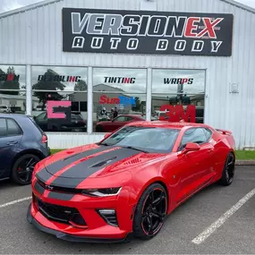 Front view of VersionEX Auto Body in Eugene, OR, showcasing a bold red Chevrolet Camaro with custom black accents. The shop exterior highlights key services including detailing, tinting, and wraps, reflecting VersionEX’s expertise in automotive customization and protection.