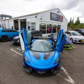 Blue McLaren with carbon accents and vertical doors open outside VersionEX Auto Body in Eugene, OR. The lineup of custom cars showcases the shop’s expertise in high-end wraps, detailing, and performance-focused automotive customization.