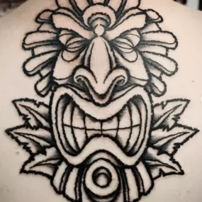 Schmierfink & Robird kräftiges traditionelles tattoo einer stilisierten maske auf dem rücken, inspiriert von polynesischen und tribal elementen, mit starken linien und kontrastreicher schattierung
