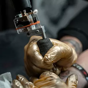 Schmierfink & Robird detailaufnahme einer tätowiermaschine während der arbeit, goldene handschuhe und präzise handhabung betonen professionalität und hygiene