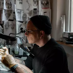 Schmierfink & Robird tätowierer im studio mit zahlreichen skizzen, arbeitet konzentriert an tattoo motiv, kreative atmosphäre und präzise handwerkskunst