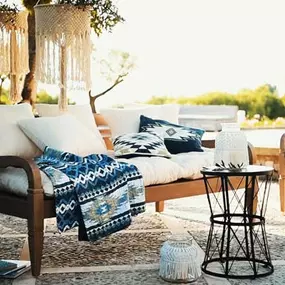 Eine stilvoll eingerichtete Terrasse mit Holzmöbeln, Textilien in Blau-Weiß und mediterranen Details lädt zum Entspannen ein. Das warme Sonnenlicht und die natürliche Umgebung erzeugen ein Gefühl von Urlaub und Leichtigkeit.