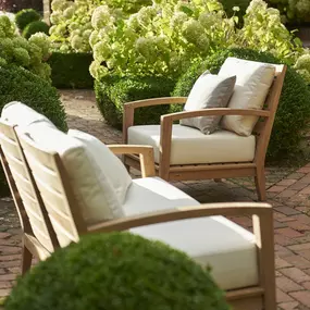 Zwei elegante Gartenstühle mit hellen Polstern stehen einladend inmitten eines gepflegten Gartens mit üppig blühenden Hortensien. Die harmonische Komposition strahlt Ruhe, Komfort und natürliche Schönheit aus.