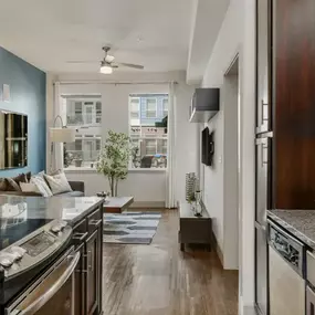 This photo showcases a stylish apartment kitchen and living room featuring dark wood cabinetry, granite countertops, stainless steel appliances, and a bright open layout. Large windows fill the space with natural light, highlighting contemporary finishes and a cozy living area with modern décor
