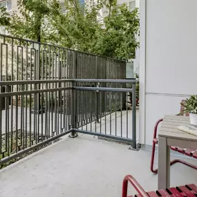 This image shows a cozy private apartment patio featuring a small table with red chairs, a potted plant, and drinks. The space is enclosed by a modern metal railing and surrounded by greenery, offering a peaceful outdoor retreat in a city setting