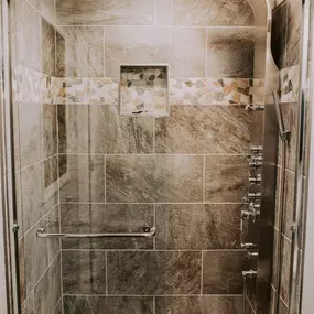 A luxurious walk-in shower featuring multi-toned stone-look tiles, a multi-jet shower panel, and a decorative pebble accent mosaic band.