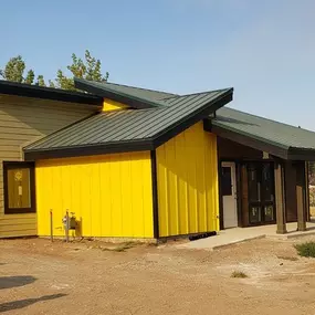 A mid-century modern ranch home with vertical tan siding and a dark green base. It features a covered porch and large bay windows.