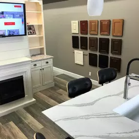 A bright kitchen and bath showroom featuring a fireplace, built-in shelving, a marbled quartz island, and a wall display of various cabinet color samples.