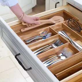 A person places wooden spoons into an open white kitchen drawer featuring a beautiful wooden cutlery and utensil organizer insert with silverware and a whisk.