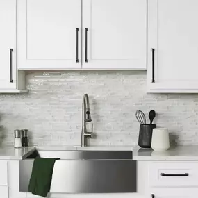A modern kitchen with white shaker cabinets, long black pulls, a stainless steel farmhouse sink, and a shimmering, light-colored glass mosaic tile backsplash.