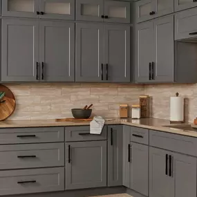 A modern kitchen featuring sleek gray shaker cabinets with black pulls, light countertops, and an earthy, horizontally laid subway tile backsplash.
