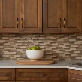 A kitchen featuring medium-toned wood cabinets with arched pulls, a bright white counter, and a narrow glass mosaic tile backsplash in natural browns and tans.