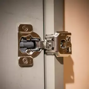 A close-up shot of an open cabinet door showing a silver-toned soft-close hinge installed on a light gray cabinet box and door.