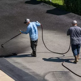 Two professional workers applying black asphalt sealcoating to a gray paved surface with spray wands. Construction Materials Asphalt & Sealcoating LLC service in Ocala, FL.