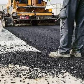 Close-up of a construction worker standing near a paving machine laying fresh, hot asphalt for a road or lot. Construction Material Asphalt & Sealcoating LLC paving services in Ocala, FL.