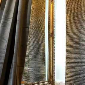 Innenraum: großer Standspiegel mit Zierrahmen, Vorhang, strukturierte Wand, schmales Tageslichtfenster und Holzfußboden.