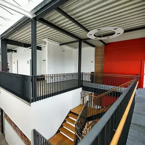 Innenraum: modernes Atrium mit Treppe, Stahlgeländer, Holzstufen, Galerie und roter Wand bei Tageslicht.