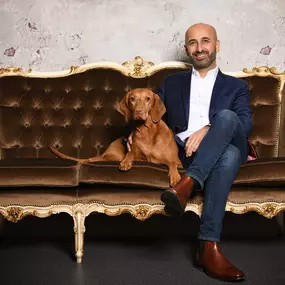 Mann im Sakko sitzt mit Hund auf barockem Samtsofa vor strukturierter Wand im Fotostudio.