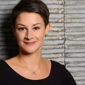 Halbportrait einer Frau mit kurzem Haar und Septum vor gerippter Metallwand; neutraler Look, direkter Blick in die Kamera.