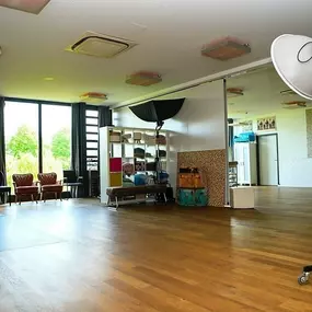 Innenraum des Fotostudios mit großer Fensterfront, Holzfußboden und mobilem Studioblitz/Softbox. Regale mit Hintergründen und Requisiten, Sitzlounge. Variable Sets für Business-, Portrait- und Familien-Shootings bei ‚ANNETTE STIER – Fotografie‘.