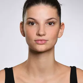 Frontaler Headshot einer jungen Frau vor hellem grauen Hintergrund; Haare zum Dutt, neutraler Ausdruck, Schultern sichtbar.