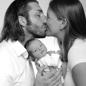 Schwarzweißaufnahme: Eltern küssen sich, halten Neugeborenes; Baby blickt in die Kamera, helles Studiohintergrund.
