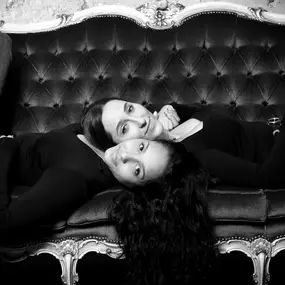 Schwarzweiß-Duo: zwei Personen liegen Kopf an Kopf auf einem barocken Samtsofa und blicken in die Kamera.